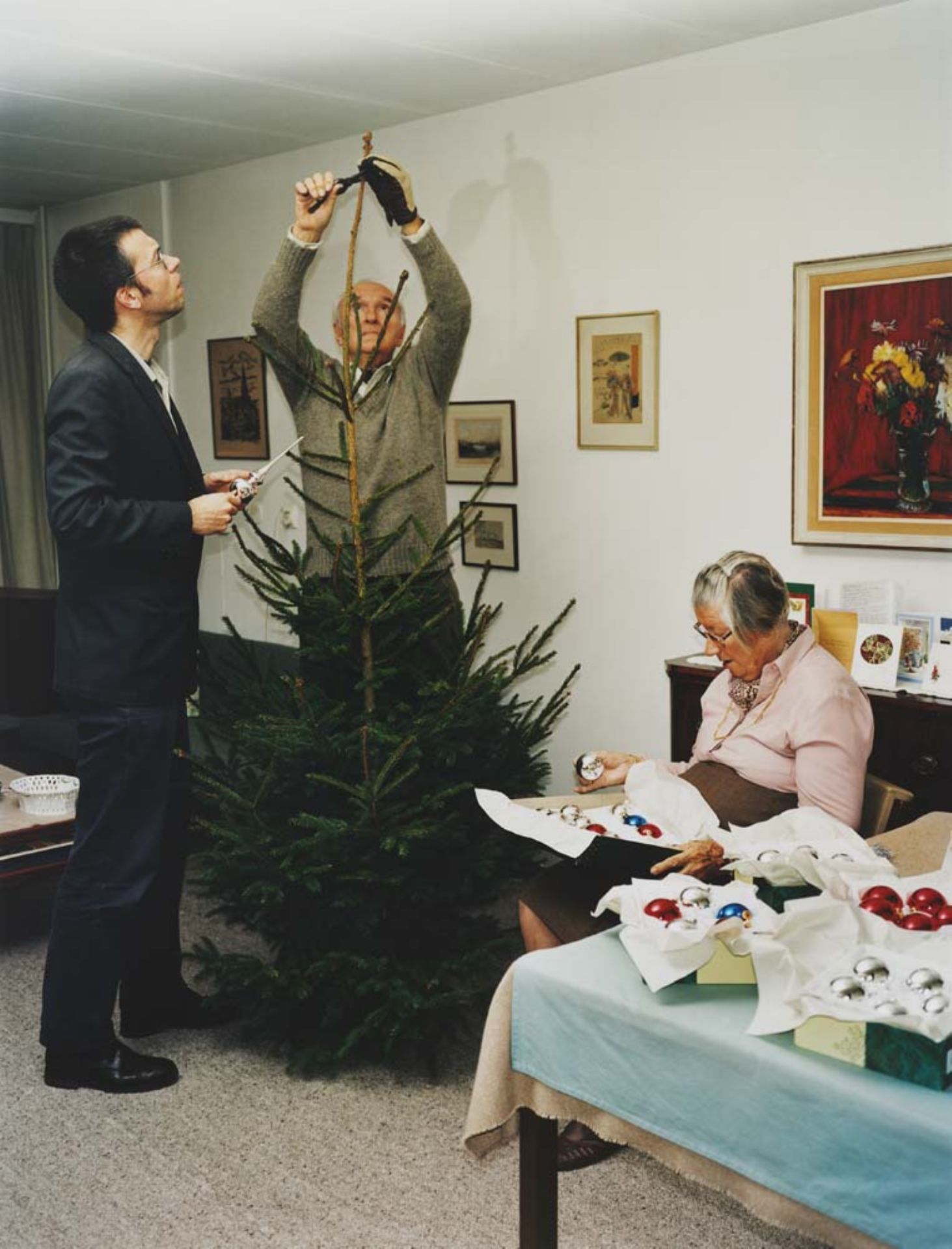 Op de foto zie je een vader, zoon en moeder in een woonkamer. Vader en zoon zijn de kerstboom aan het opzetten. Moeder is de kerstballen uit de doos aan het halen.