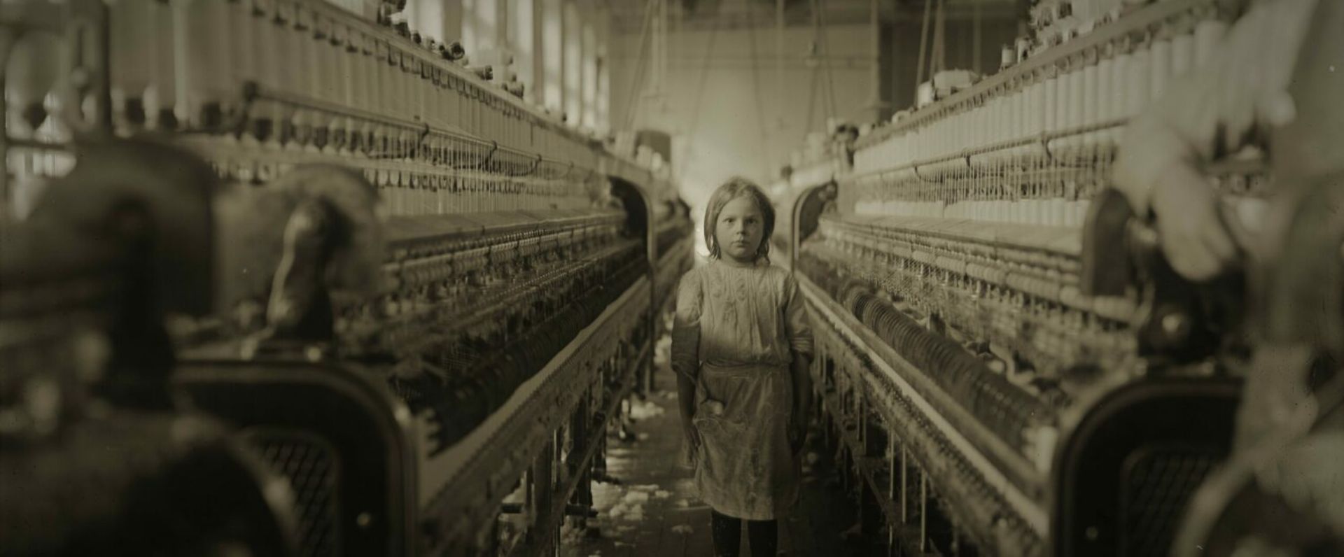 Op de foto staat een meisje of jonge vrouw tussen twee machines met daarop allemaal spoelen met garen.