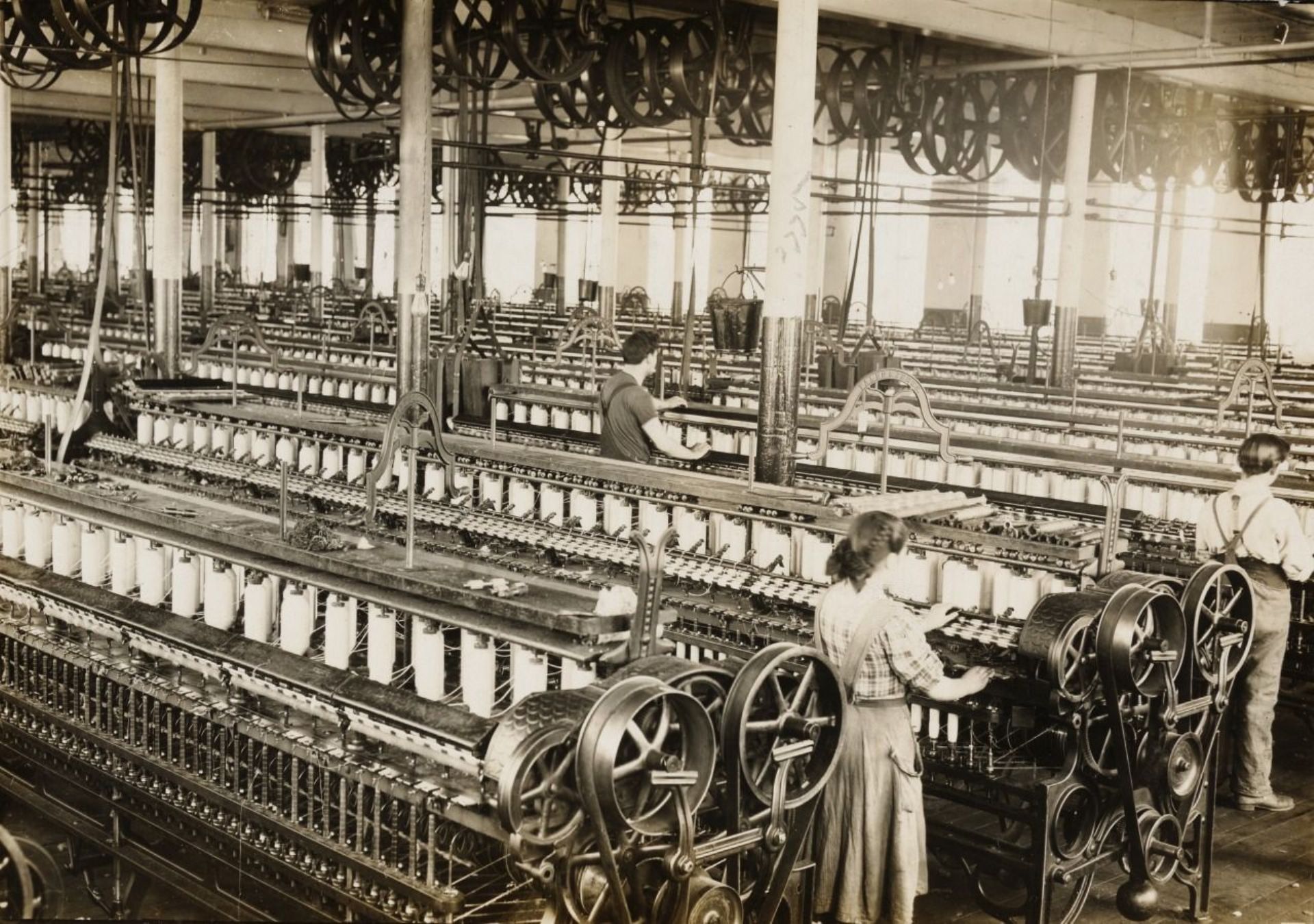 Op de foto staan veel machines waarop witte spoelen met garen staan. Er zijn ook een paar mensen aan het werk.